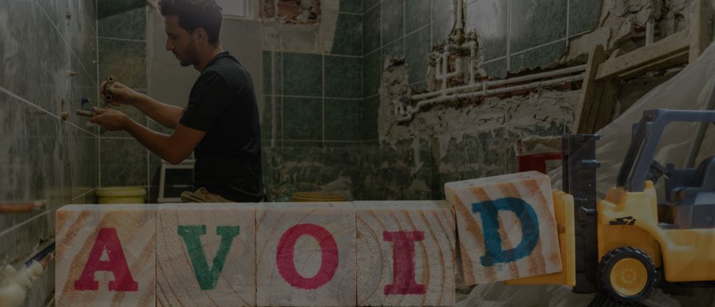 Bathroom under repair with broken tiles and a plumber redoing work, overlaid with wooden blocks spelling the word “AVOID”.